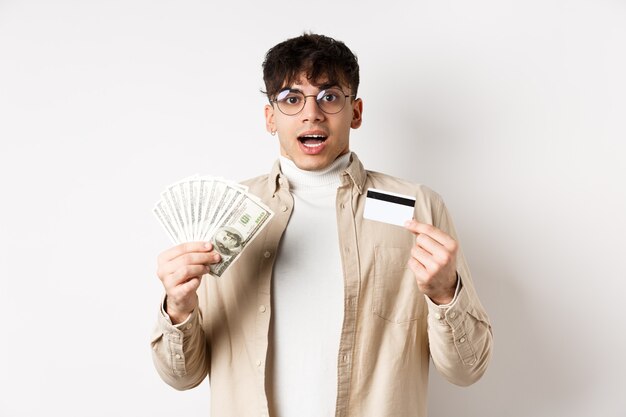 Возбужденный молодой парень показывает долларовые купюры и кредитную карту, зарабатывает деньги и выглядит изумленно, стоя на ...