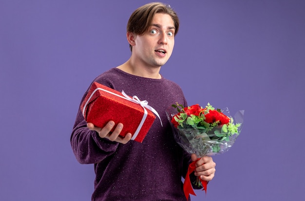 Excited young guy on valentines day holding gift box with bouquet isolated on blue background