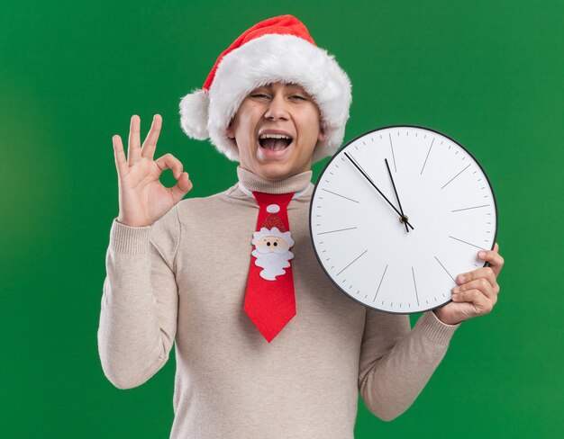 Excited young guy wearing christmas hat with tie holding wall clock showing okay gesture isolated on green wall