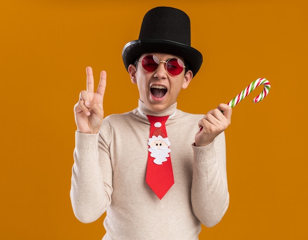 Foto gratuita eccitato giovane ragazzo che indossa il cappello con cravatta natalizia e vetri che tengono la caramella di natale che mostra gesto di pace isolato sulla parete gialla