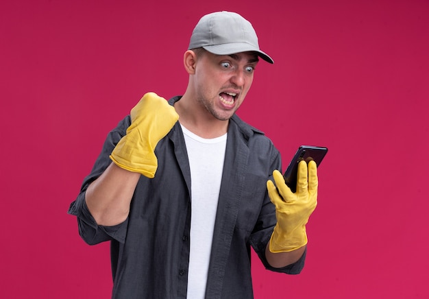 ピンクの壁に隔離されたはいジェスチャーを示している電話を保持し、見ている手袋でTシャツとキャップを身に着けている興奮した若い厄介な掃除人