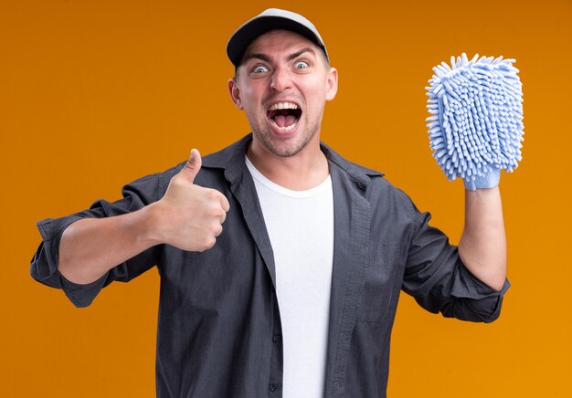 Excited young handsome cleaning guy wearing t-shirt and cap holding cleaning rag showing thumb up isolated on orange wall