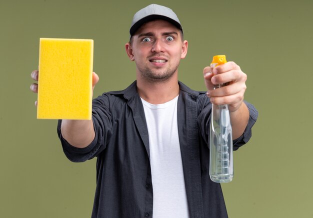 Excited young handsome cleaning guy wearing t-shirt and cap holding out sponge with spray bottle  isolated on olive green wall