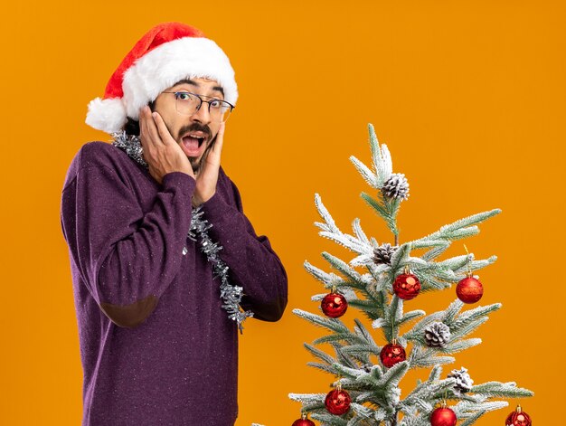 Возбужденный молодой красивый парень стоит возле елки в рождественской шапке с гирляндой на шее, положив руки на щеки, изолированные на оранжевой стене