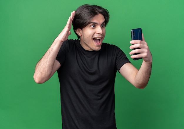 Возбужденный молодой красивый парень в черной футболке держит и смотрит на телефон, изолированный на зеленой стене