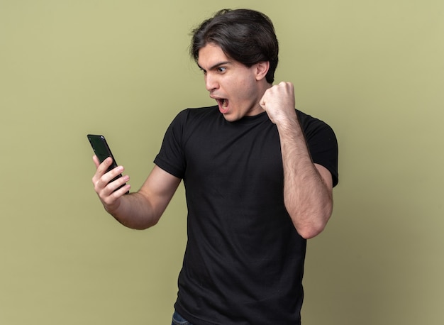 Взволнованный молодой красивый парень в черной футболке держит и смотрит в телефон, показывая жест да, изолированный на оливково-зеленой стене