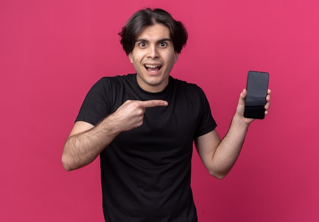 Excited young handsome guy wearing black t-shirt holding and points at phone isolated on pink wall