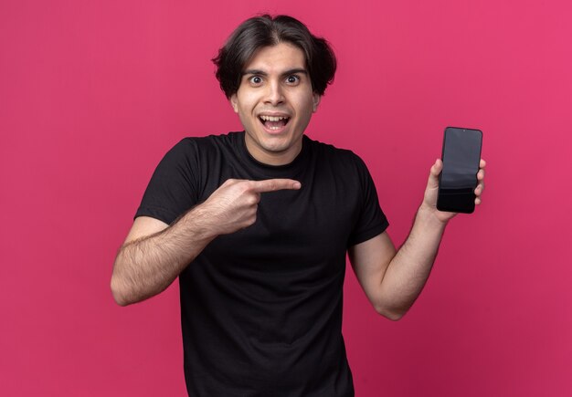 Возбужденный молодой красивый парень в черной футболке держит и указывает на телефон, изолированный на розовой стене