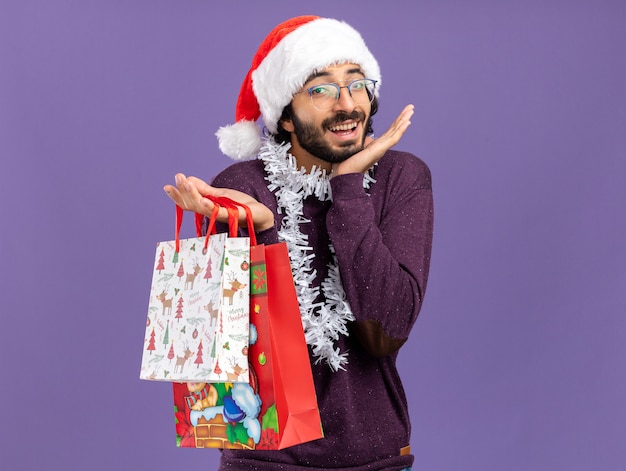 Excited young handsome guy wearing christmas hat with garland on neck holding gift bags spreading hands isolated on blue background