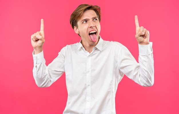 Excited young handsome guy wearing white shirt showing tongue points at up isolated on pink wall