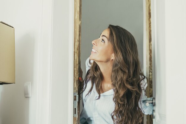 Excited young Hispanic woman coming into new apartment, opening door, standing in doorway, looking at stack of carton boxes