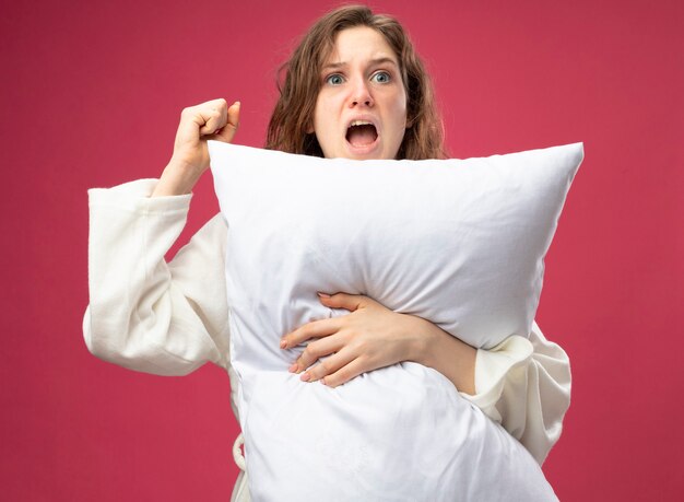 Excited young ill girl looking at side wearing white robe hugged pillow raising fist isolated on pink