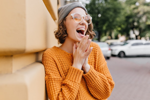 Excited young lady in gray hat laughing on the street beside building