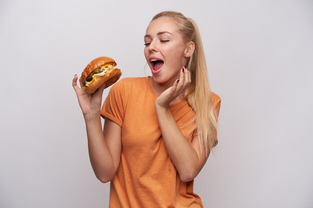 Free photo excited young lovely long haired blonde lady in orange t-shirt foretasting delicious dinner while having hamburger in raised hand, smiling pleasantly while posing over white background