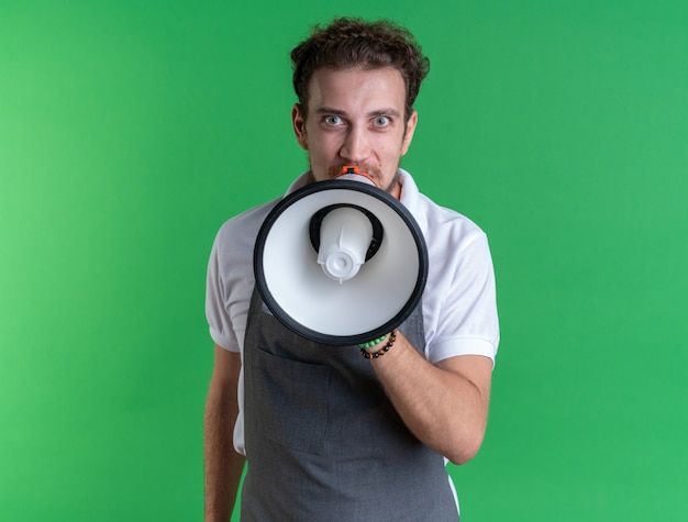 Excited young male barber wearing uniform speaks on loudspeaker isolated on green wall