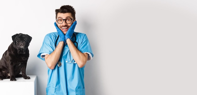 Free photo excited young male doctor veterinarian admiring cute pet sitting on table cute black pug dog waiting