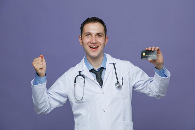 Excited young male doctor wearing medical robe and stethoscope around neck looking at camera showing credit card and fist isolated on purple background