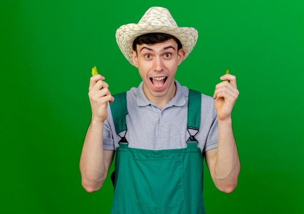 Excited young male gardener wearing gardening hat holds broken hot pepper isolated on green background with copy space