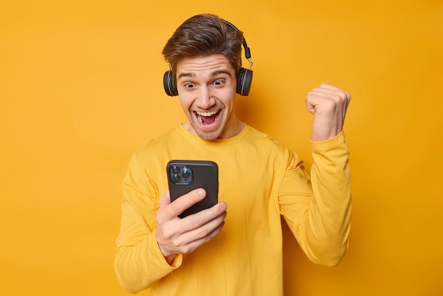 Excited young man gamer plays video game clenches fist with triumph looks at smartphone screen celebrates winning new level wears headphones on ears isolated over yellow background Yes I did it
