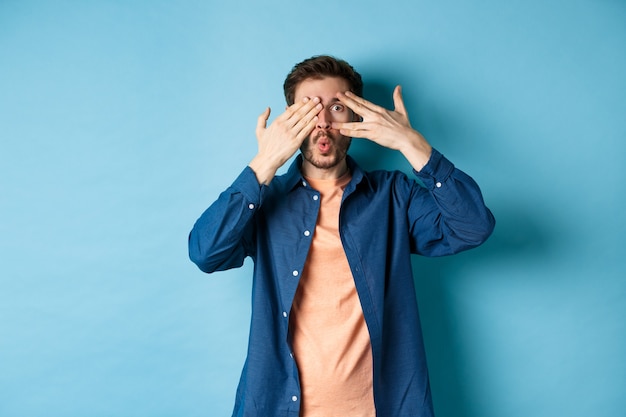 Excited young man peeking through fingers and saying wow, checking out surprise gift, standing on blue background.