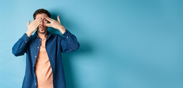 Excited young man peeking through fingers and saying wow checking out surprise gift standing on blue