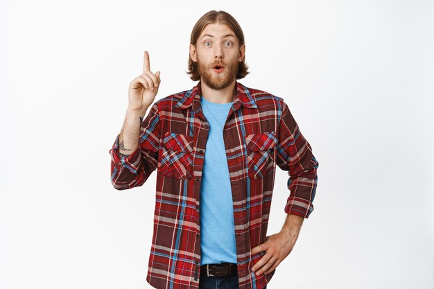 Excited young man pointing finger up, standing in casual clothes, white background.