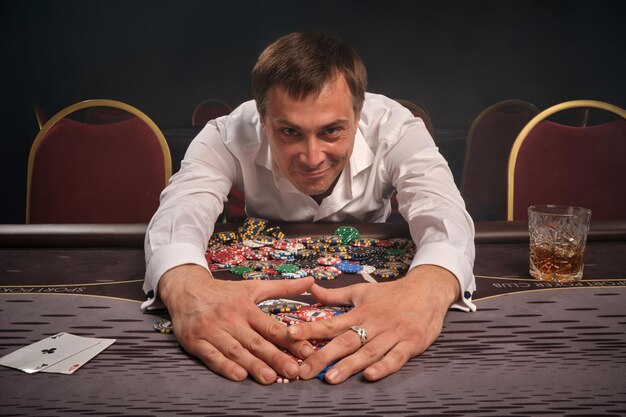 excited young man in a white shirt is playing poker sitting at the table in casino. he rejoices in winning taking the chips. gambling for money. games of fortune.
