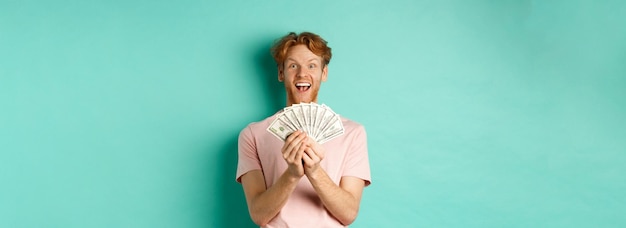 Free photo excited young man winning prize money counting cash and looking amazed at dollars standing over turq
