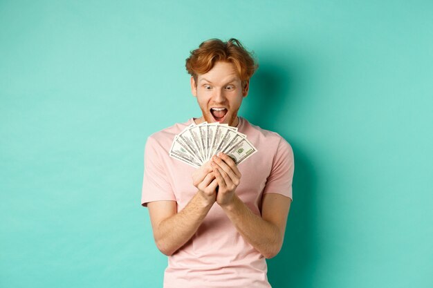상금을 받고, 현금을 세고, 청록색 배경 위에 서있는 달러에 놀란 찾고 흥분된 젊은 남자.