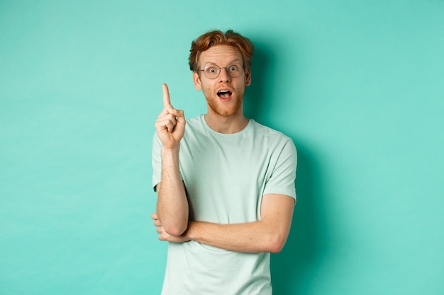 Excited young man with ginger hair in glasses, raising index finger, pitching an idea, standing over mint background