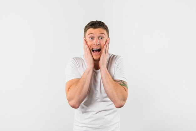 Excited young man with his two hands on cheeks looking to camera against white backdrop