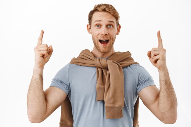 Excited young man with red hair big strong body pointing fingers up and smiling amazed give advice recommend to click link showing shopping deal standing over white background