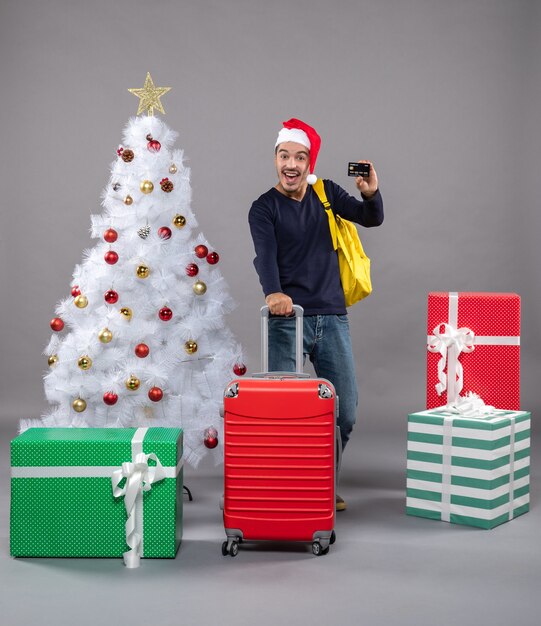 灰色のカードを示す赤いスーツケースを保持しているサンタ帽子を持つ興奮した若い男