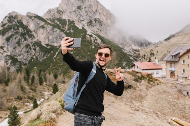 Excited young man with short haircut posing in mountains with cozy house
