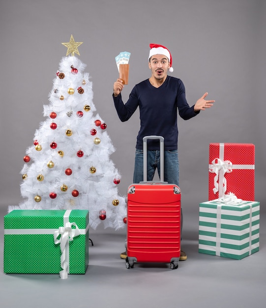 クリスマスツリーの周りに旅行チケットと赤いスーツケースと灰色のプレゼントで興奮した若い男