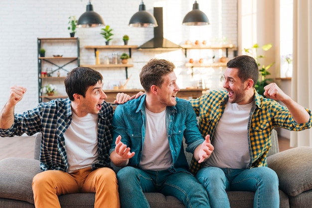 Excited young men sitting on sofa cheering at home