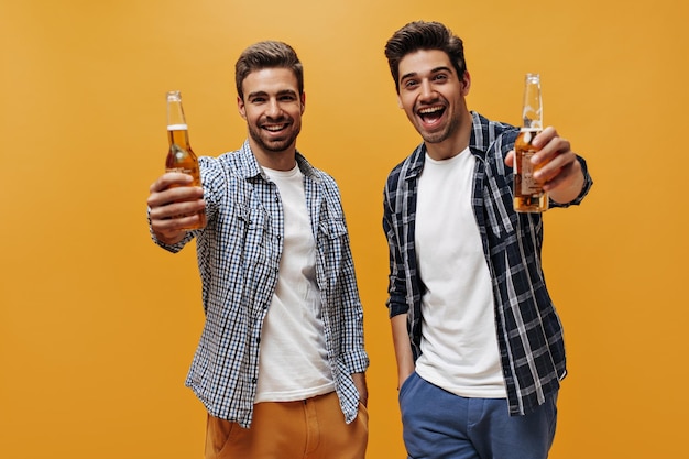 Excited young men in white trendy tshirts and checkered shirts rejoice and hold beer bottles on orange isolated background