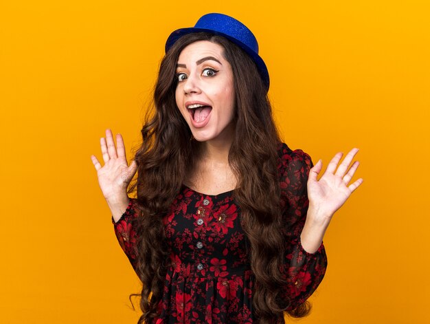 Excited young party woman wearing party hat looking at front showing empty hands isolated on orange wall