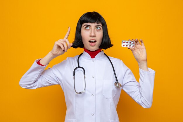 Возбужденная молодая симпатичная кавказская девушка в униформе врача со стетоскопом держит упаковку таблеток и указывает вверх