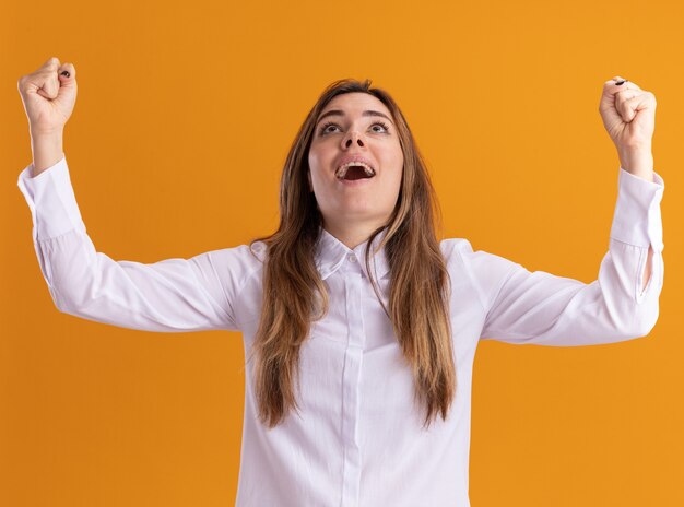 Excited young pretty caucasian girl stands with raised fists and looks up isolated on orange wall with copy space