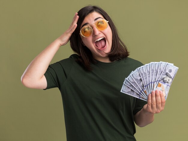 Excited young pretty caucasian girl in sun glasses puts hand on head and holds money looking at side