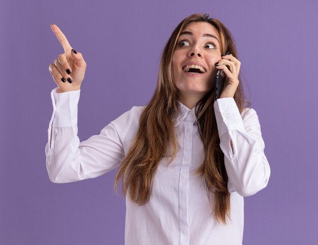 Excited young pretty caucasian girl talks on phone looking and pointing at side