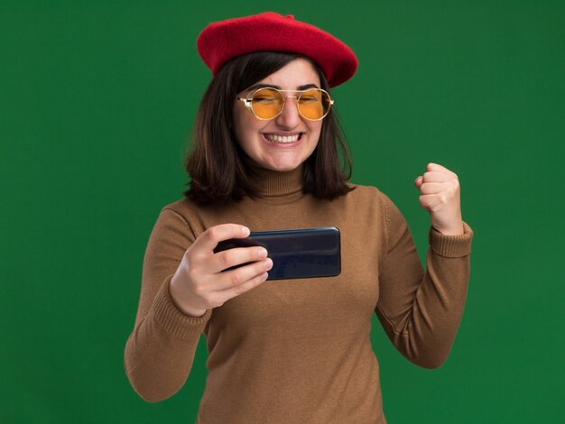 Excited young pretty caucasian girl with beret hat in sun glasses keeps fist and holds phone