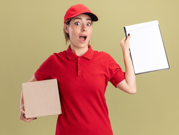 Возбужденная молодая симпатичная доставщица в униформе держит буфер обмена и картонную коробку на оливково-зеленом