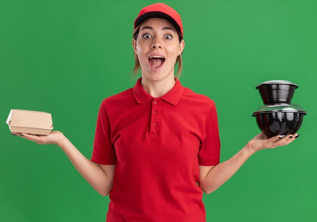 La giovane ragazza graziosa di consegna emozionante in uniforme tiene i contenitori di cibo e il pacchetto di cibo sul verde