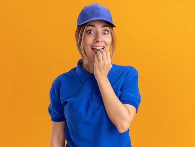 Excited young pretty delivery girl in uniform puts hand on mouth on orange