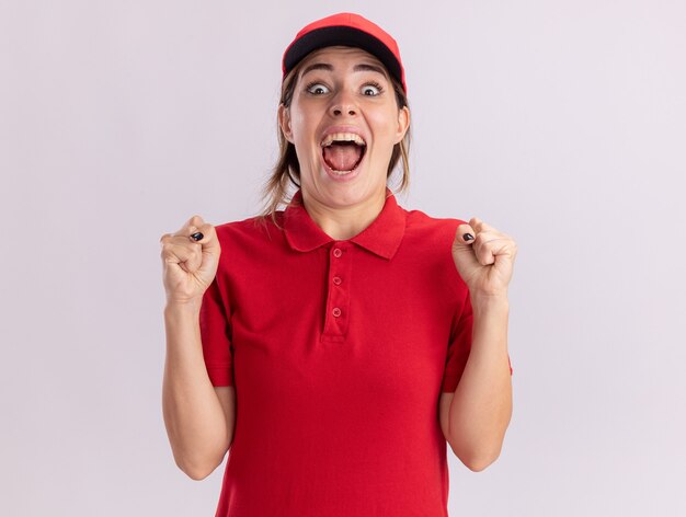 Excited young pretty delivery woman in uniform keeps fists isolated on white wall