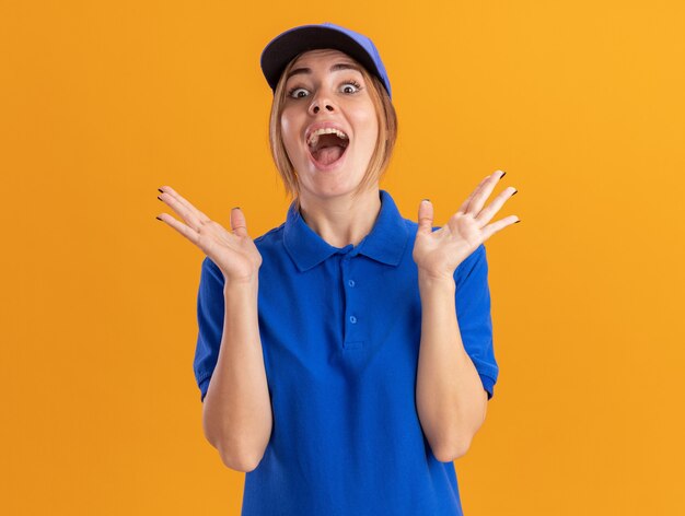 Excited young pretty delivery woman in uniform stands with raised hands isolated