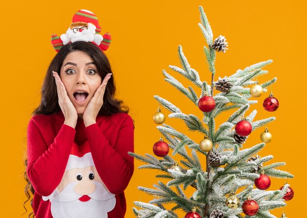 오렌지 배경에 고립 된 카메라를보고 얼굴에 손을 유지 장식 된 크리스마스 트리 근처에 산타 클로스 머리띠와 스웨터 서 입고 흥분된 젊은 예쁜 여자