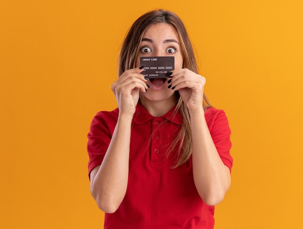 Бесплатное фото Возбужденная молодая красивая женщина держит кредитную карту, изолированную на оранжевой стене